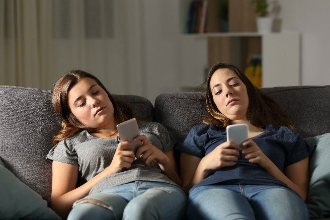 Social-Media-Stagnation-Bored-People two women on a couch looking at their cell phones.