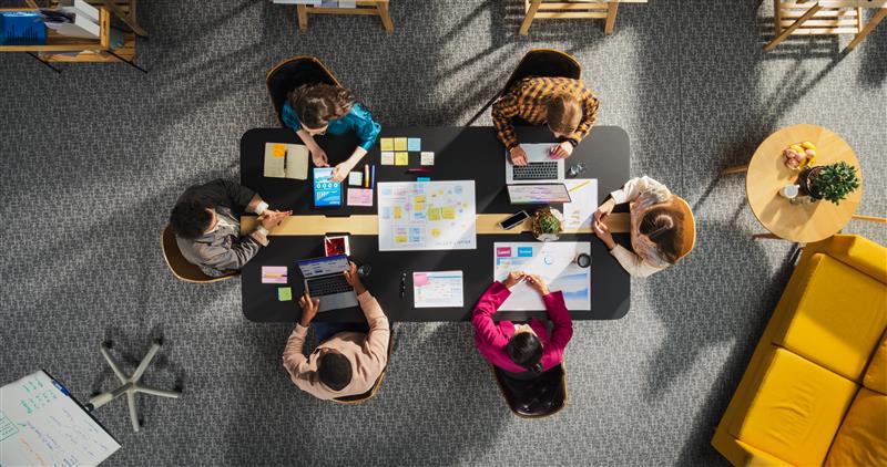 Team collaborating around a table with laptops, charts, and notes in a modern office setting.
