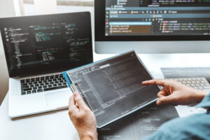 A person holding a tablet, with a laptop and monitor screen in the background. All three screens show code being developed.